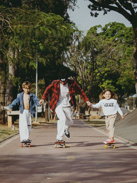 Pai e filhos andam de skate lado a lado, de mãos dadas, por um caminho entre árvores.