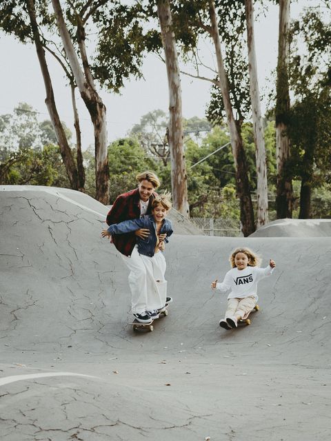 O pai, de camisa xadrez vermelha, desce a rampa de uma pista de skate abraçando um dos filhos, que ri; a filha vem atrás, sentada no skate, de braços abertos.