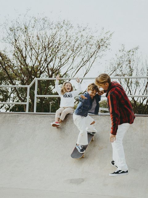 Na rampa de skate, a menina levanta os braços, sorrindo, enquanto o pai se curva para ajudar o menino a ficar em pé sobre o skate.