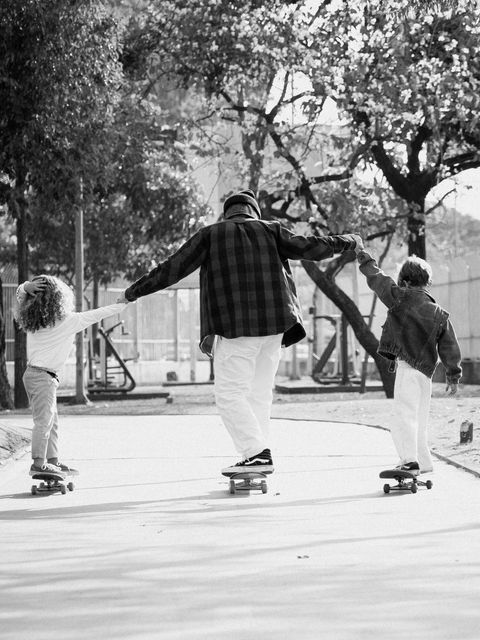 Em preto e branco, o pai e os dois filhos, vistos de costas, andam de skate de mãos dadas por um caminho no parque.