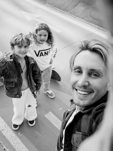 Em preto e branco, selfie do pai sorrindo para a câmera, com os dois filhos ao lado, de pé na pista.