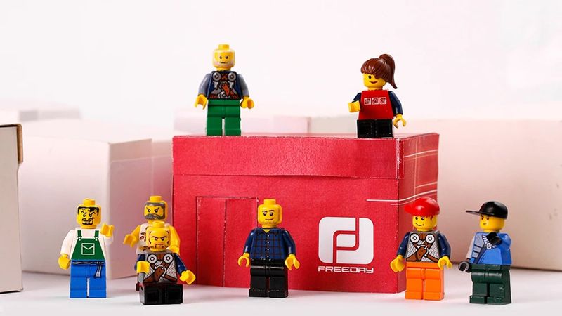Quadro do vídeo: bonecos de Lego em volta de uma caixa de tênis vermelha com o logotipo da Freeday, entre caixas brancas.