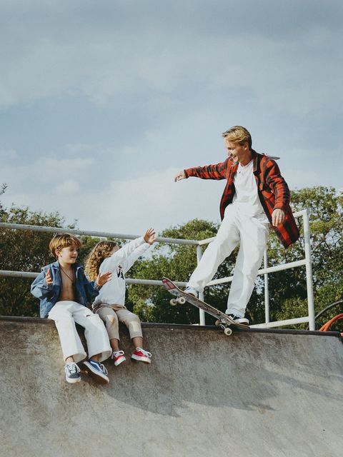 O pai desliza o skate na borda de uma pista com o braço estendido, enquanto os dois filhos o observam sentados na grade, com árvores e céu ao fundo.